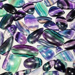 Puede incluir: Una colección de piedras preciosas de fluorita pulidas en varias formas, incluyendo ovaladas, en forma de lágrima y rectangulares. Las piedras muestran una gama de colores, incluyendo púrpura, verde, azul y blanco, con patrones arremolinados. Las piedras están dispuestas sobre una superficie blanca.
