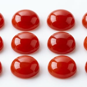 May include: Twelve polished, cabochon-cut, red gemstones arranged in three rows of four. The stones have a smooth, rounded surface and a glossy finish, reflecting light. The gemstones are a vibrant, solid red color.