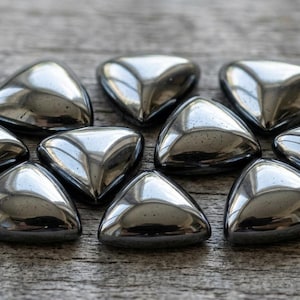 May include: A collection of ten polished, triangular hematite stones. Each stone has a smooth, reflective surface, appearing dark gray with metallic highlights. They are arranged on a weathered, light brown wooden surface.