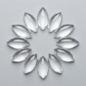 Puede incluir: Doce cabujones de vidrio transparente e incoloro, con forma de marquesa, dispuestos en un patrón circular similar a una flor. Cada pieza presenta una superficie lisa y abovedada con extremos puntiagudos, creando una exhibición simétrica.