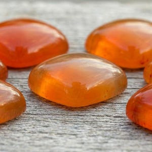 Può includere: Primo piano di sette pietre preziose arancioni e lucide. Le pietre sono lisce, arrotondate e traslucide, con un aspetto caldo e luminoso. Sono disposte su una superficie di legno grigio usurato. Le pietre preziose sono probabilmente utilizzate per gioielli o scopi decorativi.