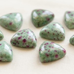 Peut inclure: Huit pierres précieuses triangulaires polies, avec un motif vert et rouge moucheté. Les pierres sont disposées sur une surface beige clair, mettant en valeur leur texture lisse et brillante. Idéal pour la joaillerie ou la collection.