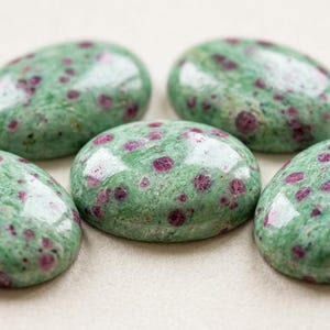 May include: Five oval-shaped gemstones with a polished finish. The stones have a green base color with small, irregular red inclusions. The gemstones are arranged in a circular pattern, showcasing their natural beauty.