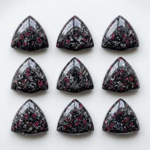 Puede incluir: Nueve cabujones triangulares pulidos dispuestos en una cuadrícula de tres por tres. Cada piedra presenta una base negra oscura con inclusiones moteadas grises y blancas y puntos de color rojo magenta vibrante en la superficie lisa y brillante.
