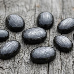 May include: Nine polished, oval-shaped black stones with a reflective surface are arranged on a weathered, gray wooden surface. The stones have a smooth, glossy appearance, and the wood grain is visible.