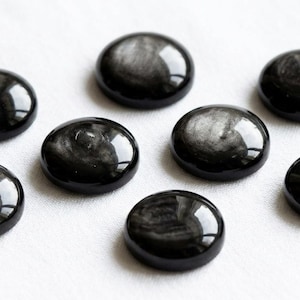 Peut inclure: Huit cabochons en obsidienne noire polis, de forme ovale, avec une surface brillante et réfléchissante. Chaque pierre présente un motif argenté tourbillonnant à l'intérieur, sur fond sombre. Les pierres sont disposées sur une surface blanche.