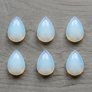 Peut inclure: Dix cabochons lisses et translucides en forme de goutte, avec une finition irisée blanc laiteux et bleu pâle. Les pierres sont disposées en deux rangées horizontales de cinq, chacune présentant une base arrondie et un sommet pointu.