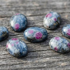 May include: Seven oval-shaped cabochon gemstones featuring a polished surface with a mottled blue and grey base color, interspersed with distinct, vibrant pink inclusions and small metallic flecks throughout each individual stone.