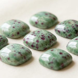 May include: Eight polished, square-shaped gemstones. The stones are a mottled green color with small, dark red inclusions. The gemstones are arranged in a circular pattern on a light beige surface.