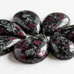 May include: A collection of six polished, teardrop-shaped cabochons featuring a dark black base with speckled grey inclusions and vibrant, rounded magenta-coloured spots distributed across the smooth, reflective surfaces of each individual stone.