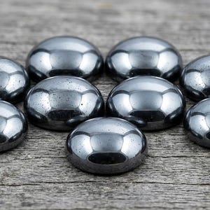 May include: Nine polished, gray, cabochon gemstones arranged on a weathered wooden surface. The stones have a smooth, rounded dome shape and reflect light, creating highlights. The gemstones are likely used for jewelry making or other crafts.