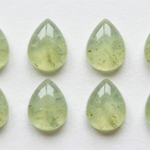 May include: Eight pear-shaped prehnite gemstones. The translucent stones have a pale green hue with darker green inclusions. The polished gems are arranged in two rows of four on a white surface, showcasing their natural beauty.