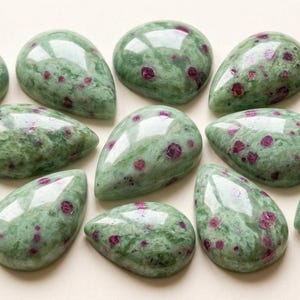 May include: A collection of teardrop-shaped gemstones. The stones are a mottled green colour with small, irregular red inclusions. The polished surfaces reflect light, and the stones are arranged on a light-coloured surface.