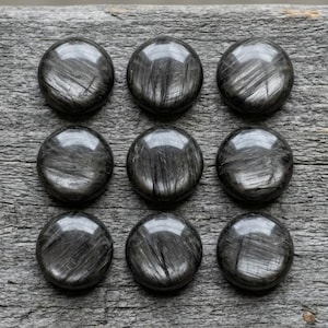 May include: Nine round, polished, black stones with a reflective surface and a linear pattern are arranged in a 3x3 grid on a weathered, gray wooden surface. The stones have a subtle sheen.