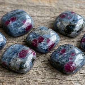 May include: Seven polished, cushion-shaped kyanite gemstones featuring a smooth, glossy surface. Each stone displays a marbled blue and grey base colour with distinct, vibrant magenta-red ruby inclusions scattered across the crystalline structure.
