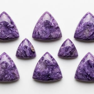 May include: Eight triangular, polished charoite gemstones. The stones vary in size and are arranged on a white surface. The gemstones display a swirling pattern of deep purple and lavender hues, with some areas of translucent clarity.