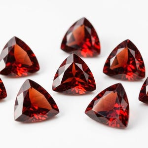 Könnte beinhalten: Acht dreieckige, facettierte, tiefrote Edelsteine. Sie sind auf einer weißen Oberfläche angeordnet und zeigen ihren Brillantschliff und ihre satte Farbe. Diese Edelsteine sind wahrscheinlich Granate, ideal für die Schmuckherstellung.