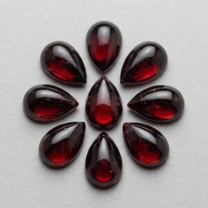 Könnte beinhalten: Eine Ansammlung von neun tiefroten, birnenförmigen Granat-Edelsteinen, die in einem kreisförmigen, blumenartigen Muster angeordnet sind. Jeder Stein hat eine glatte, polierte Cabochon-Oberfläche, die das Licht reflektiert und einen satten, dunkelroten Farbton zeigt.
