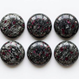Puede incluir: Diez cabujones circulares dispuestos en dos filas de cinco. Cada cabujón presenta una superficie pulida y lisa con una base moteada en gris oscuro y negro, intercalada con distintas inclusiones minerales de color rojo magenta.