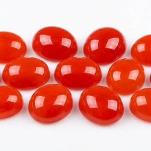 May include: A collection of eleven polished, oval-shaped, vibrant red gemstones. Each stone has a smooth, glossy surface, reflecting light. The gemstones are arranged on a white background, showcasing their color and form.