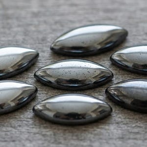 May include: Seven polished, oval-shaped, dark gray stones arranged in a circular pattern. The stones have a smooth, reflective surface, and are set against a textured, light brown wooden background. The stones appear to be hematite.