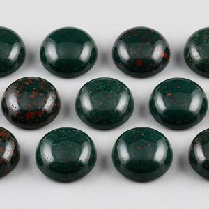May include: A collection of twelve round, polished gemstones. The stones are a deep, forest green with speckled red inclusions. The cabochons are arranged in three rows of four, each with a smooth, domed surface.