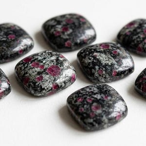 Op de afbeelding: Zeven gepolijste, vierkante cabochons met een gevlekte zwart-grijze stenen basis met ingebedde levendige magenta-roze cirkelvormige insluitingen. Elk stuk heeft afgeronde randen en een gladde, reflecterende textuur over het hele oppervlak.