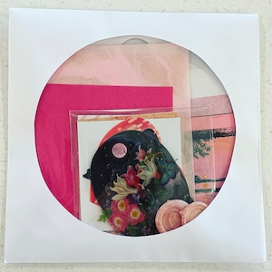May include: A collection of art supplies, including pink and fuchsia paper, stickers, and a decorative seal, presented in a white frame with a circular opening. The stickers feature floral and celestial designs.