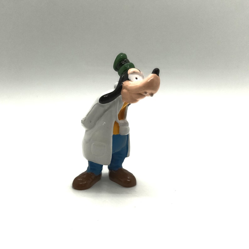Pode incluir: Uma pequena e colorida figura do Pateta, um personagem de desenho animado da Disney. Pateta est&aacute; vestindo um jaleco branco, cal&ccedil;as azuis, sapatos marrons e um chap&eacute;u verde. Seu grande focinho bronzeado &eacute; proeminente e ele parece estar olhando para baixo.