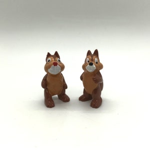 Pode incluir: Duas pequenas figuras de esquilos de desenho animado, castanhos e bege. Os esquilos t&ecirc;m rostos brancos, narizes vermelhos e orelhas castanhas escuras. Est&atilde;o em p&eacute; com os bra&ccedil;os ao lado. O fundo &eacute; branco liso.