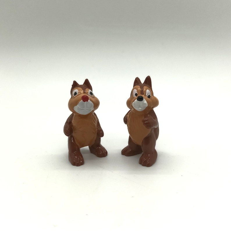 Pode incluir: Duas pequenas figuras de esquilos de desenho animado, castanhos e bege. Os esquilos t&ecirc;m rostos brancos, narizes vermelhos e orelhas castanhas escuras. Est&atilde;o em p&eacute; com os bra&ccedil;os ao lado. O fundo &eacute; branco liso.