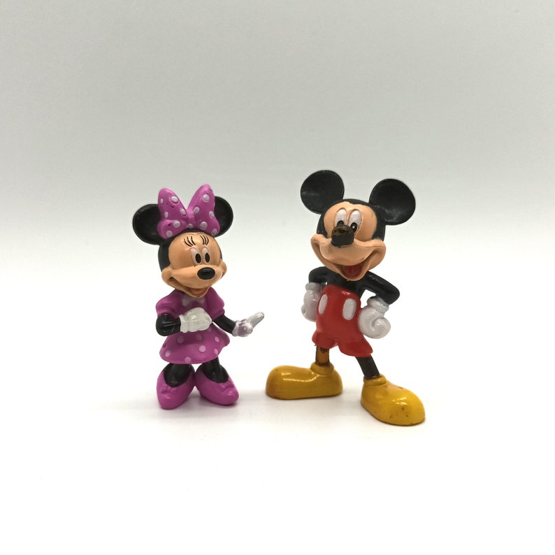 Pode incluir: Duas figuras dos personagens da Disney Mickey Mouse e Minnie Mouse. Mickey Mouse est&aacute; vestindo shorts vermelhos, sapatos amarelos e luvas brancas. Minnie Mouse est&aacute; vestindo um vestido rosa com bolinhas brancas, um la&ccedil;o combinando e sapatos rosa.