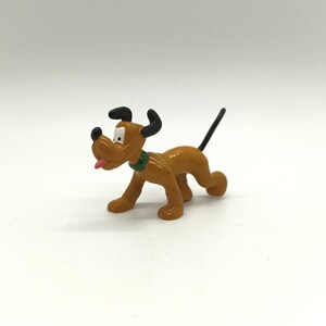 Pode incluir: Uma pequena figura de Pluto marrom com orelhas pretas e uma cauda preta. O c&atilde;o tem uma coleira verde e uma l&iacute;ngua rosa. A figura est&aacute; em posi&ccedil;&atilde;o de caminhada.