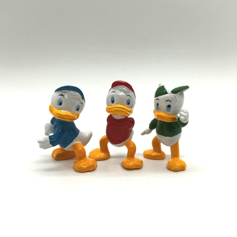 Pode incluir: Tr&ecirc;s estatuetas do Pato Donald em v&aacute;rias poses. Cada figura tem um corpo branco, bico e p&eacute;s amarelos e uma roupa de cor diferente: azul, vermelho e verde. As estatuetas est&atilde;o em uma superf&iacute;cie branca.