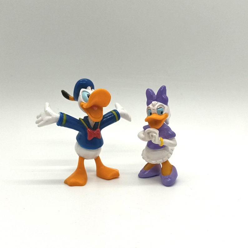 Pode incluir: Duas figuras da Disney do Pato Donald e da Margarida. Pato Donald usa um traje e chap&eacute;u de marinheiro azuis, com bico e p&eacute;s laranja. Margarida usa top e sapatos roxos, com vestido e la&ccedil;o brancos. Ambos est&atilde;o em p&eacute;.