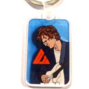 May include: A blue and orange keychain featuring a cartoon illustration of a person playing a guitar. The keychain is rectangular and has a silver ring.