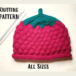 May include: A knitted strawberry hat in bright pink with a green top. The hat has a textured pattern and the words "KNITTING PATTERN" and "ALL SIZES" are displayed. Two silver knitting needles are next to the hat.