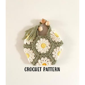 Könnte beinhalten: Eine Häkeltasche mit einem Gänseblümchenmuster in Weiß und Grün. Die Tasche hat einen Holzgriff und einen Quasten.