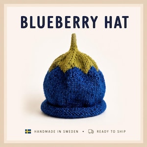 May include: A knitted hat shaped like a blueberry with a blue base and a green top featuring a small stem. The text BLUEBERRY HAT is printed in dark blue at the top. The hat has a textured knit pattern and a rolled brim at the bottom edge.