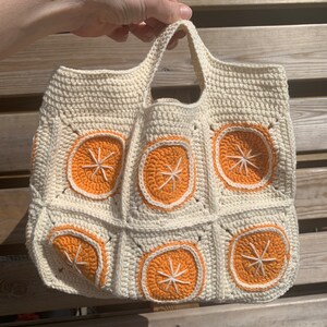 May include: Cream-colored crochet tote bag featuring orange slice designs. The bag has a square shape with a handle and is made of a textured yarn. The orange slices are outlined in white and have white starburst centers.