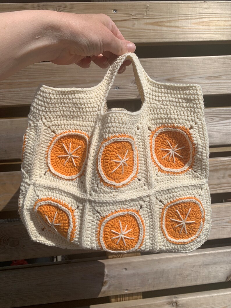 May include: Cream-colored crochet tote bag featuring orange slice designs. The bag has a square shape with a handle and is made of a textured yarn. The orange slices are outlined in white and have white starburst centers.