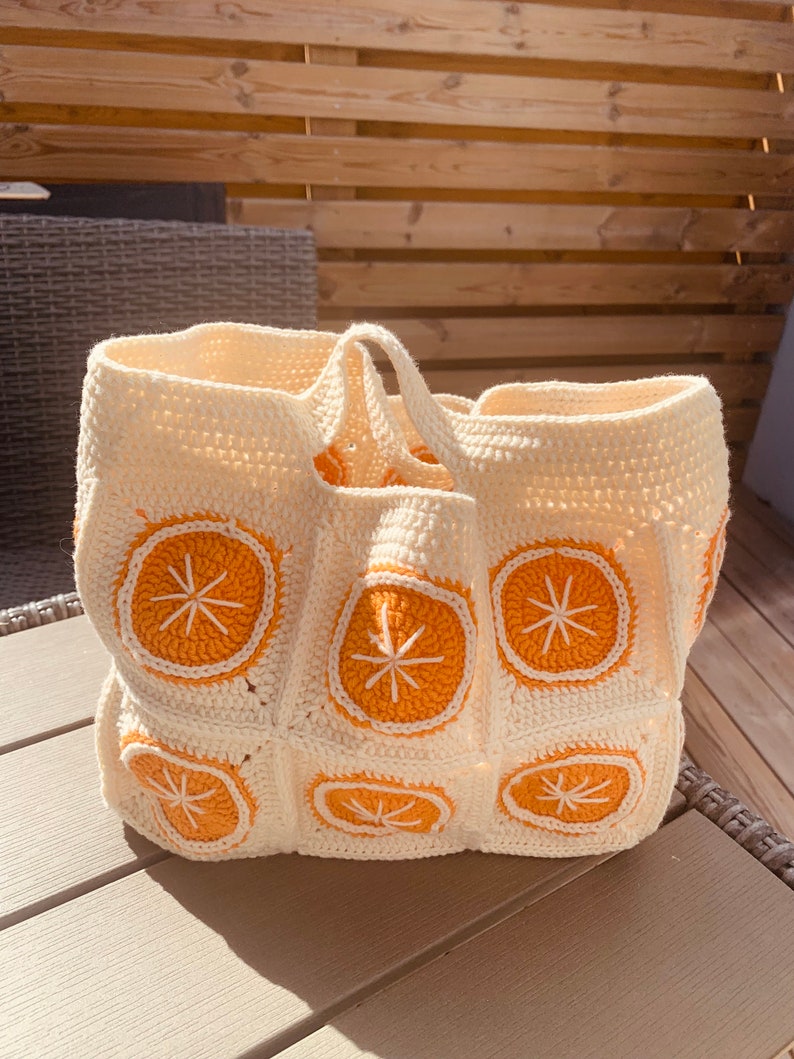 May include: Cream-colored crochet tote bag with orange citrus slice designs. The bag features a rectangular base and two handles. The orange slices have white embroidered details. The bag is sitting on a wooden surface.