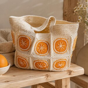 May include: A cream-colored crochet tote bag with orange citrus slice designs. The bag is made of a textured yarn and has a rectangular shape with a handle. An orange sits in a small white bowl on a wooden surface.