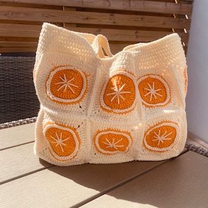May include: Cream-colored crochet tote bag featuring orange citrus slice designs. The bag has a rectangular shape with a wide opening and two handles. The orange slices have white details. A handmade, unique accessory.