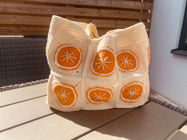 May include: Cream-colored crochet tote bag featuring orange citrus slice designs. The bag has a rectangular shape with a wide opening and two handles. The orange slices have white details. A handmade, unique accessory.