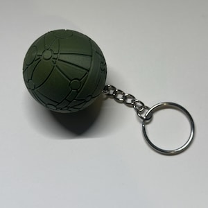 May include: A dark green, spherical keychain charm with a detailed geometric pattern. The charm is attached to a silver chain and a keyring. The design features a series of interconnected lines and circular shapes.