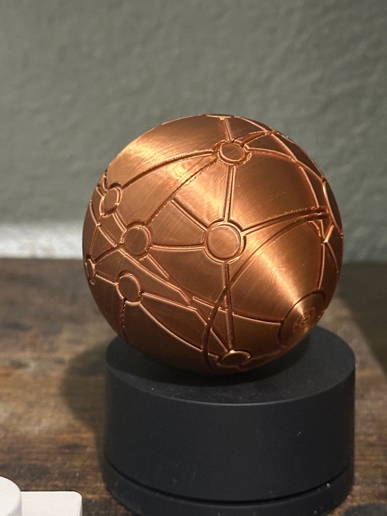 May include: A copper-coloured sphere with a geometric pattern of lines and circles. The sphere is on a black circular base.