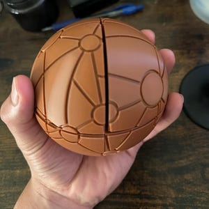May include: A spherical puzzle object in a matte brown finish, featuring a complex network of raised lines and circular nodes across its surface. The sphere is divided into two distinct halves by a vertical seam running through the centre.