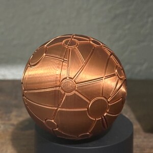 May include: A copper-coloured sphere with a geometric pattern of lines and circles. The sphere is sitting on a black base.