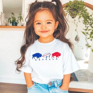 May include: A white t-shirt featuring a graphic of three buffalo in blue, white, and red, with the word "BUFFALO" below. The shirt is worn by a young child wearing blue jeans.